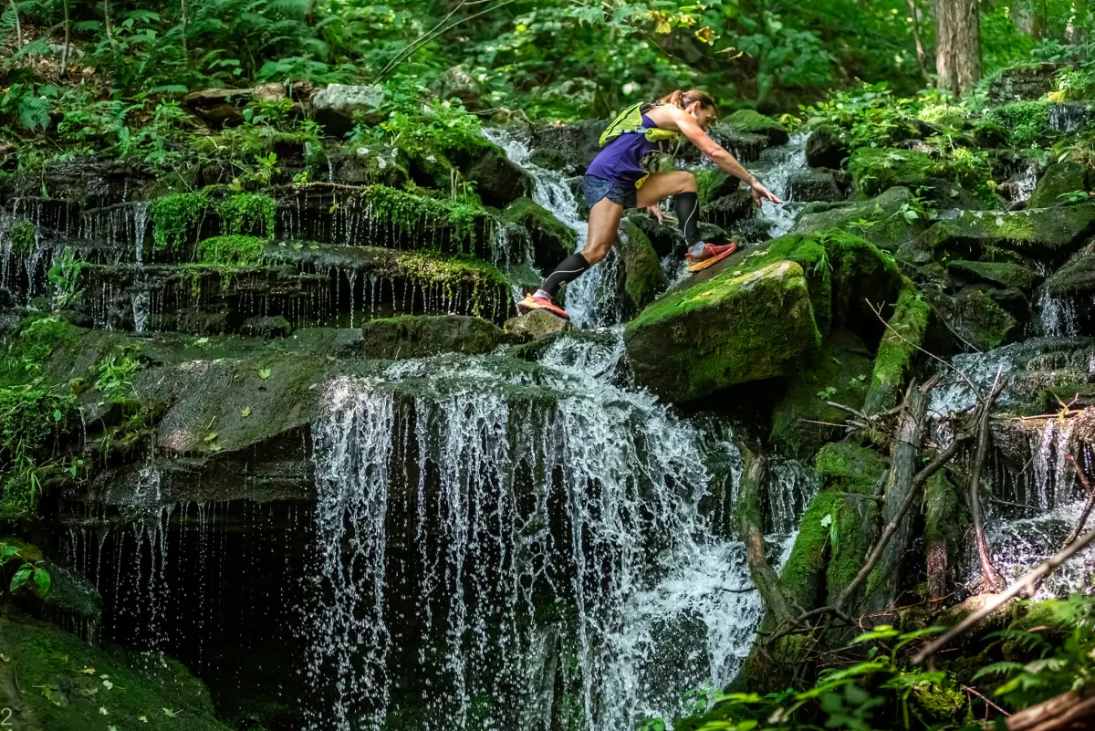 A trail runner climbing over mossy rocks beside a waterfall at Blue Knob
